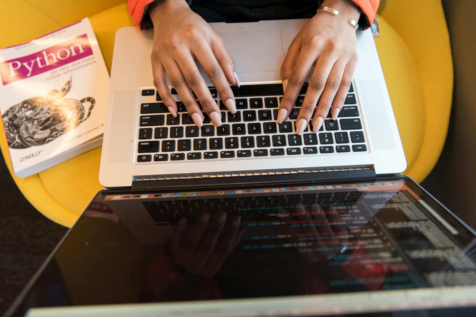 Close-up of hands typing on a laptop keyboard with a Python programming book in sight